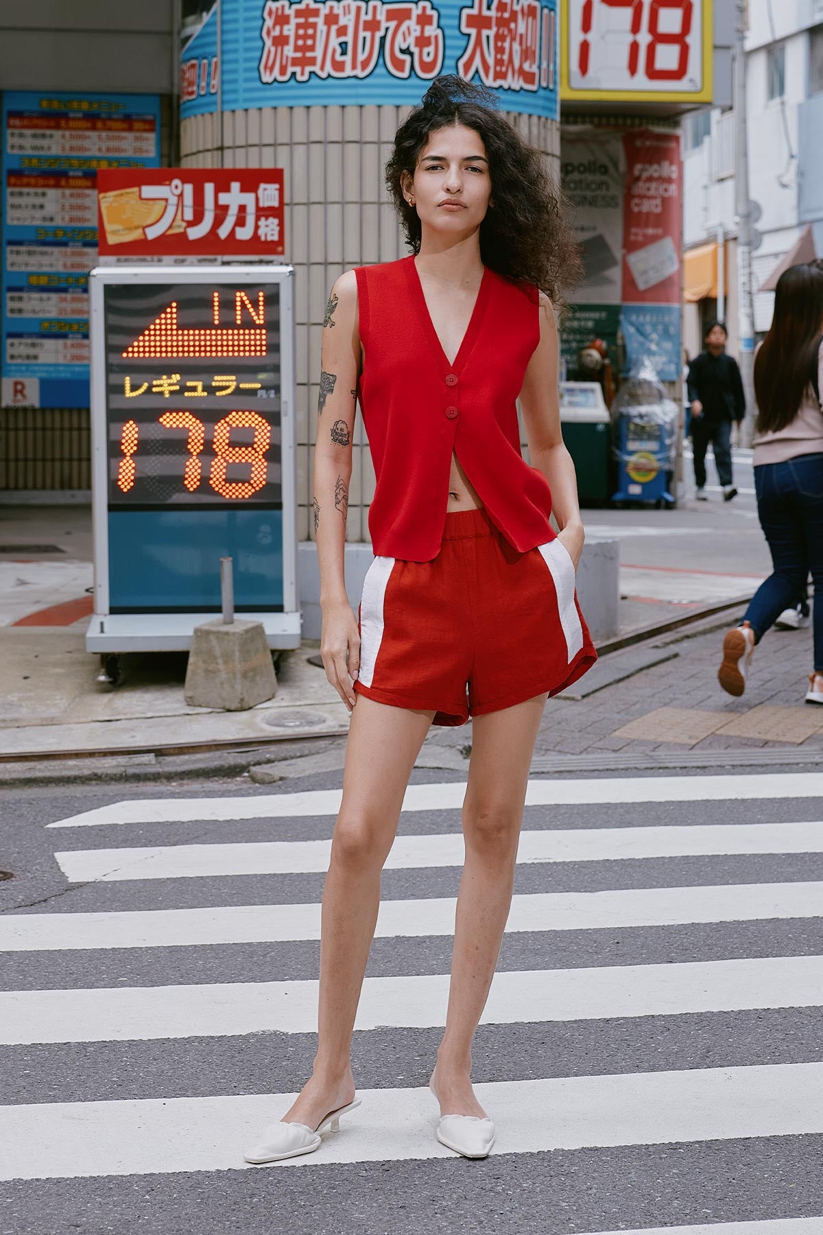 Woman in a red linen shorts by ROWIE The Label, paired with a red Goldie Knit Vest standing on a crosswalk in Tokyo, Japan with various signs and advertisements in the background.