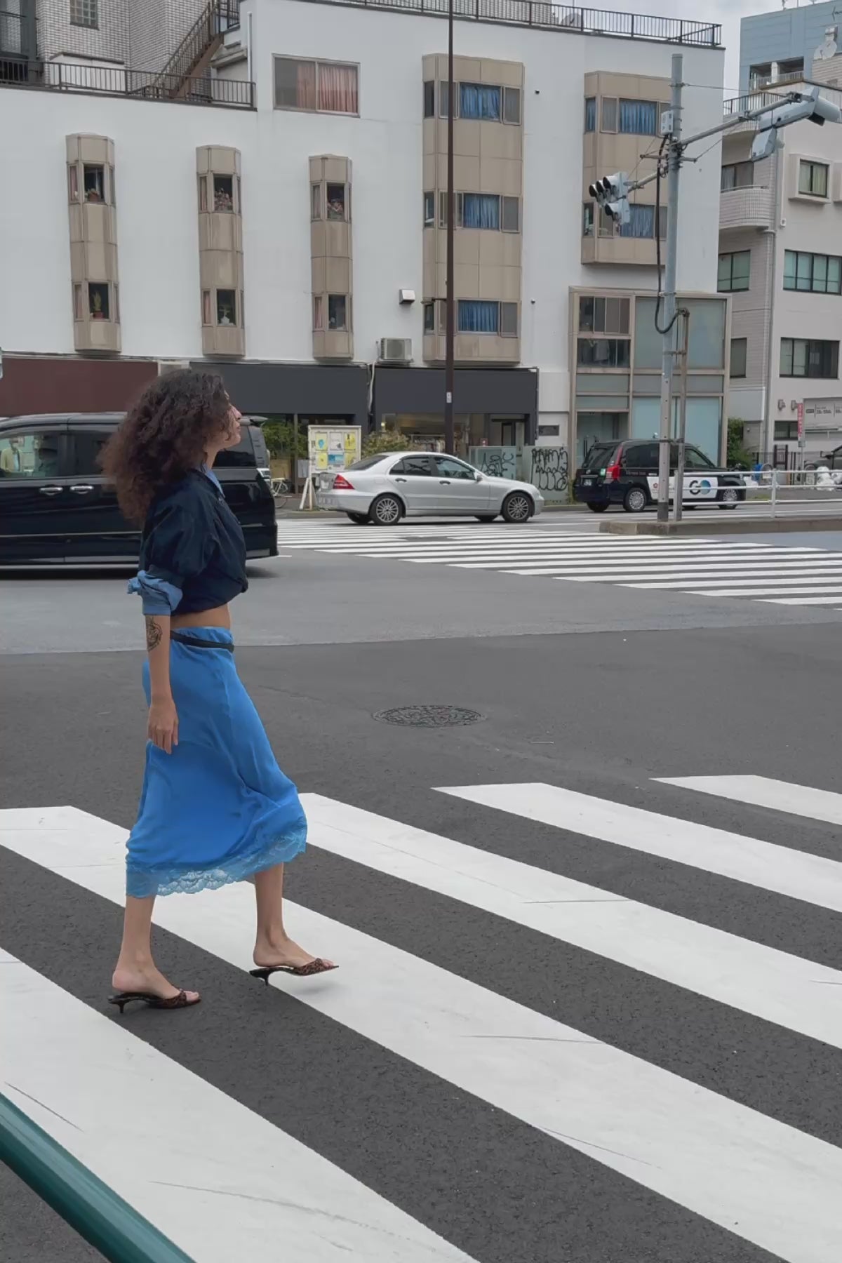 Video of model wearing a Marina Silk Midi Skirt in Cornflower, styled with a Ramona Silk Long Sleeve Shirt in Periwinkle layered under a Cora Spring Blouse in Ink Cotton by ROWIE the Label, filmed on a Tokyo street.