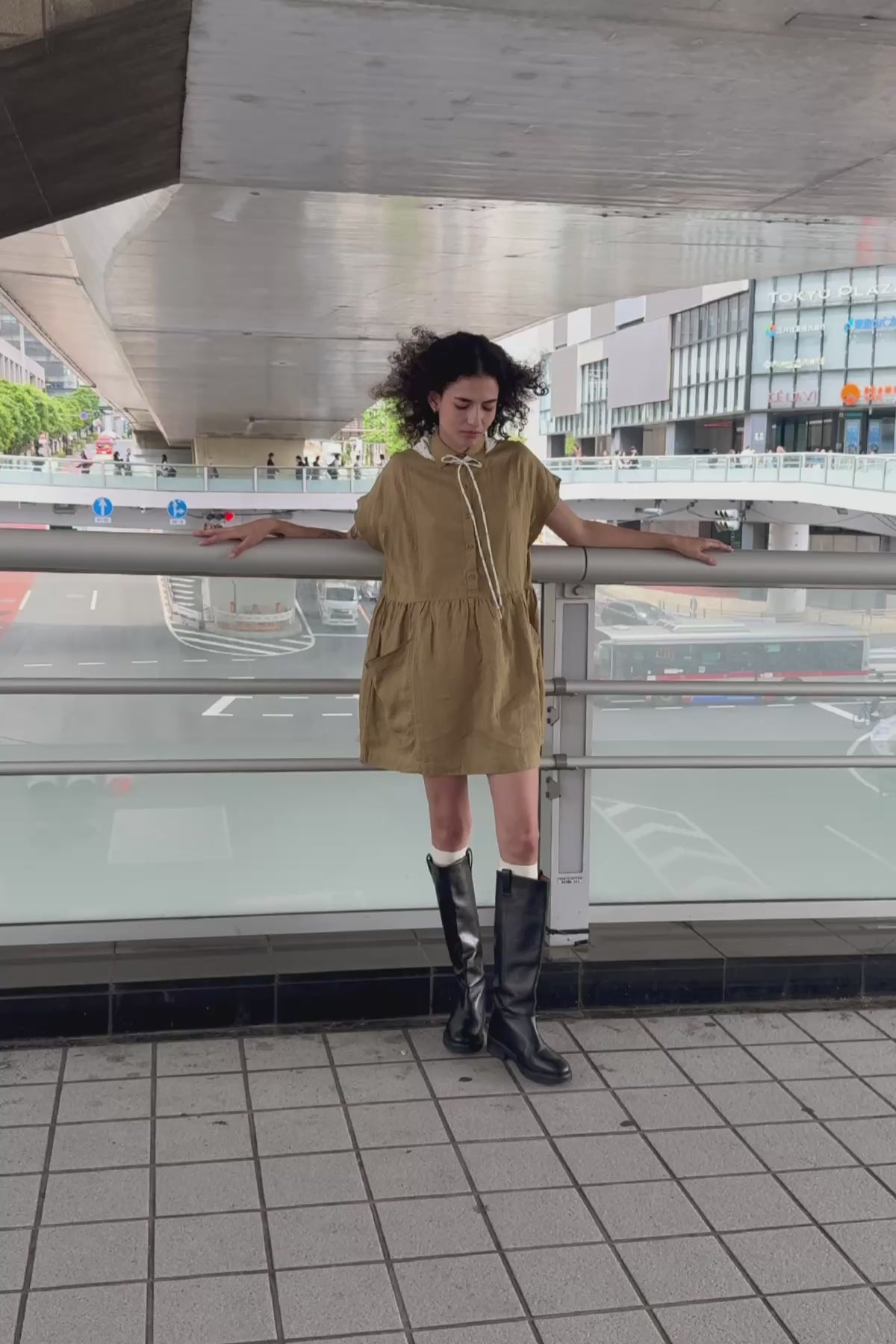 Video of model wearing a Gina Linen Mini Dress in Dusty Olive by ROWIE the Label, styled with a Frannie Silk Belt Scarf in Vanilla Spot tied around her neck, standing in an urban outdoor setting in Tokyo.