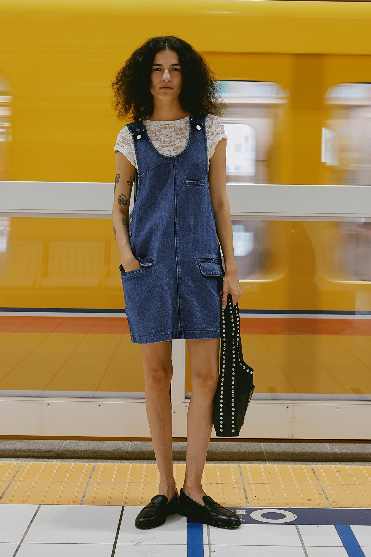 Model in Franka Denim Mini Dress with a White Galo Lace Tee underneath, standing on a subway platform with a yellow train in the background.