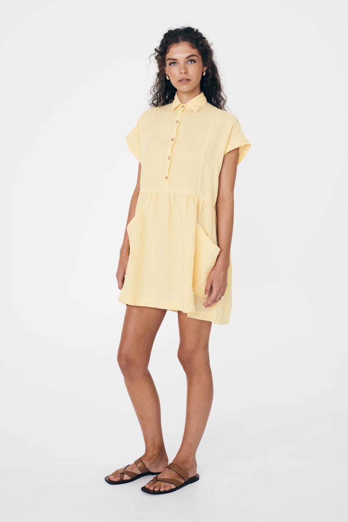 Woman wearing a light yellow, button up,  linen dress with large pockets and brown sandals