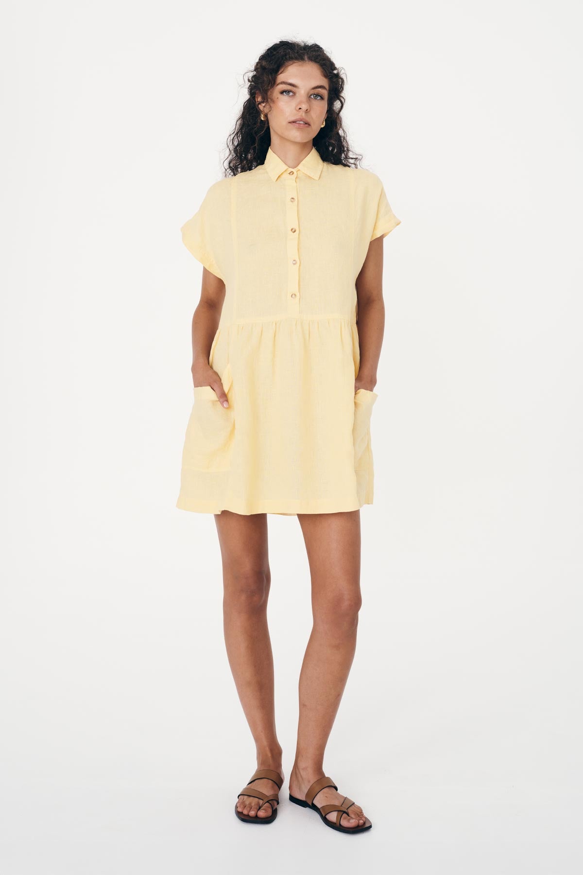 Model wearing a ROWIE The Label french linen, summer dress in lemon yellow. The Gina Linen Mini Dress buttons up to the collar with pockets on either side 