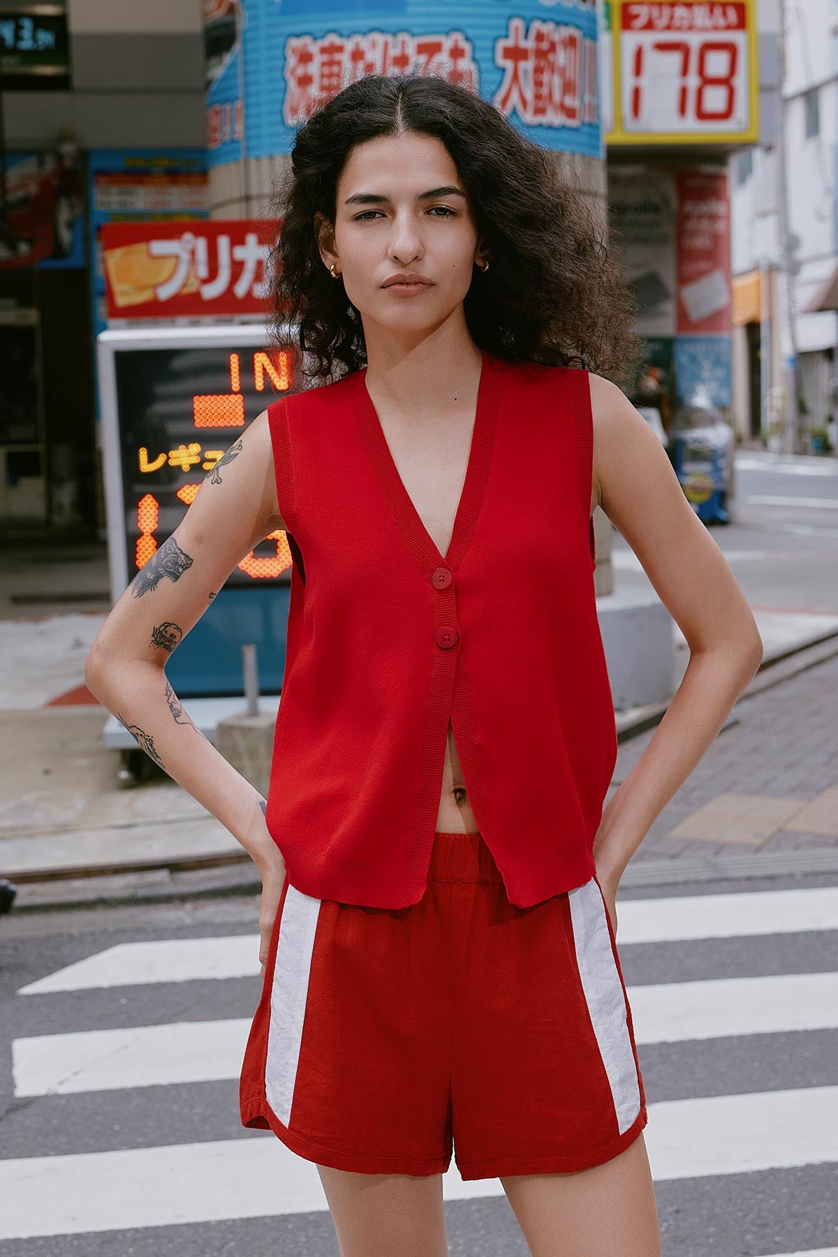 Model wears a Goldie Knit Vest in Siren Red, paired with red Linen Shorts and set again Tokyo city backdrop