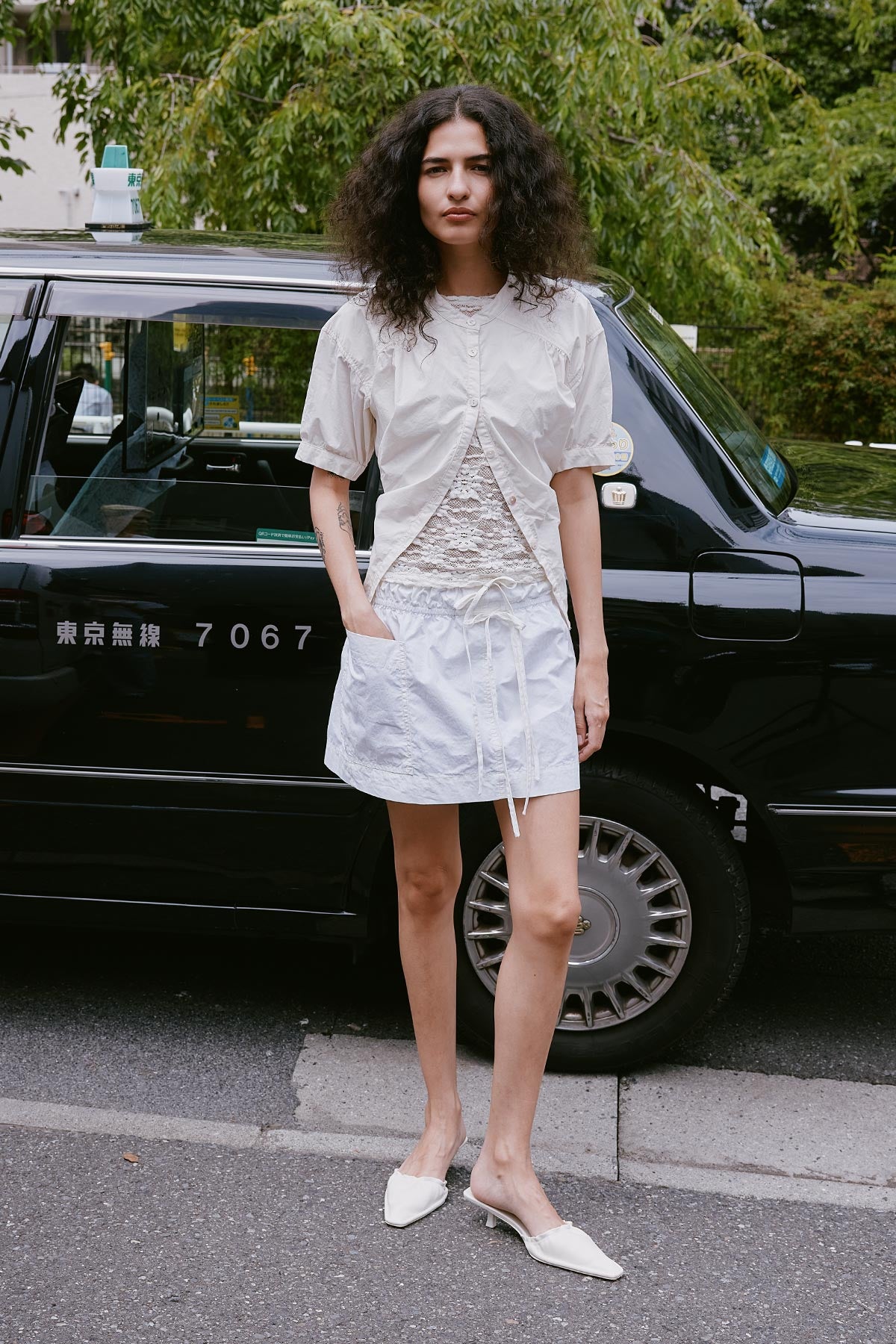 Model stands in front of black taxi in Japan wearing a ROWIE The Label Jessica Cargo Mini Skirt in white, paired with a Galo Sunflower Lace Tee Shirt and a Cora cropped white blouse over the top