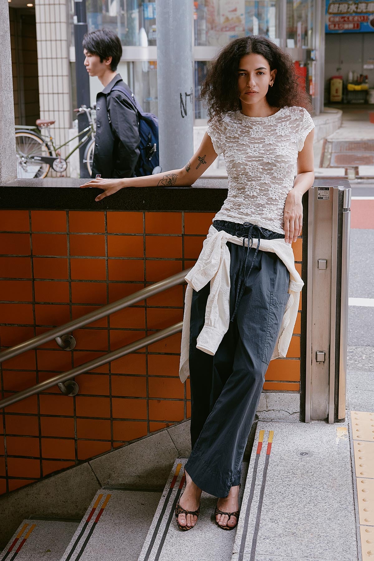 Woman stands on subway entrance in Tokyo, Japan wearing Jessica Cargo Pants by ROWIE The Label paired with the Galo Lace Tee, with sunflower lace design and the Ramona Silk Shirt tied around her waist