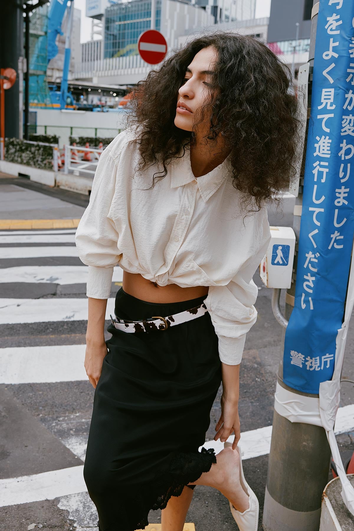 Woman walking on a street in Tokyo wearing a Ramona Silk Shirt, tied at the waist and paired with a ROWIE The Label Marina Silk Skirt in Black