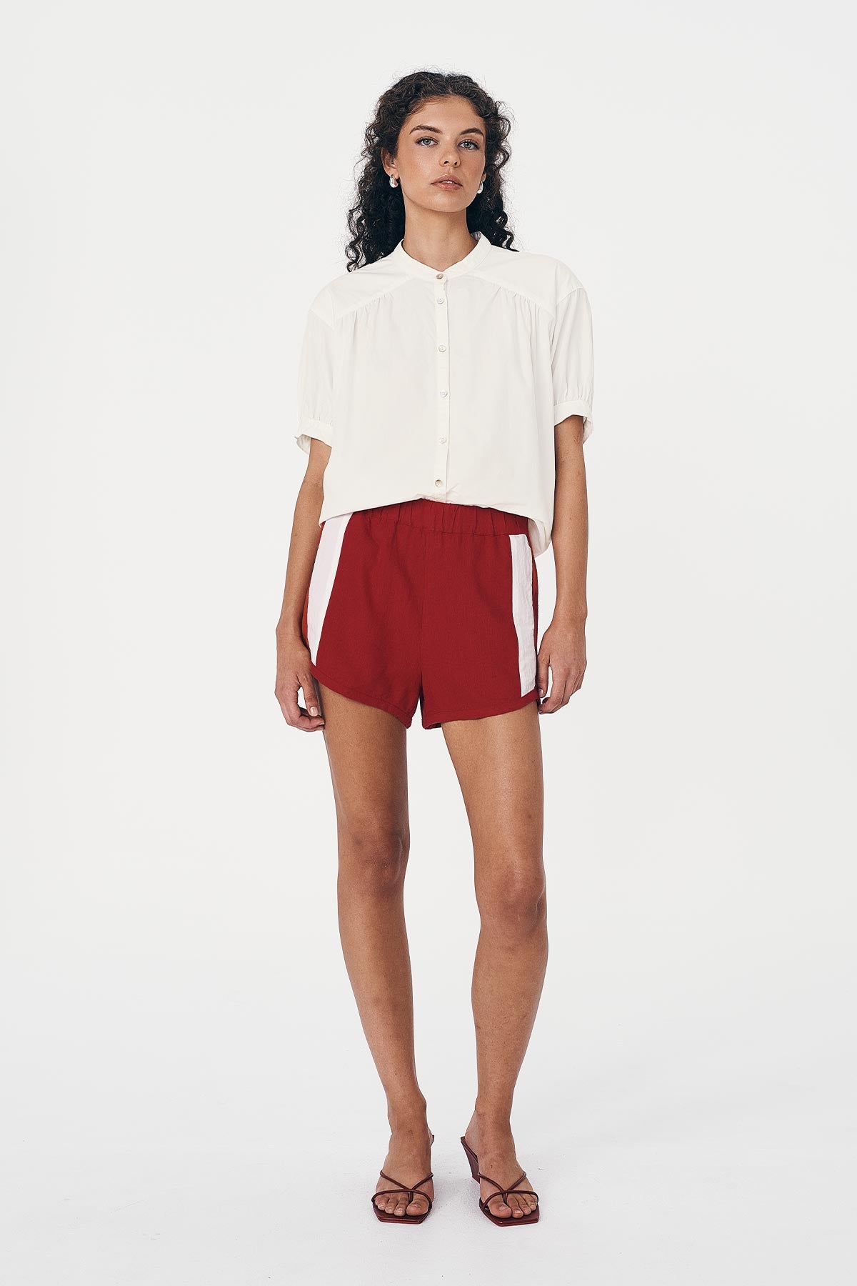 Woman wearing a white Cora Spring Blouse and red Simone Linen Shorts with a white stripe on a white background