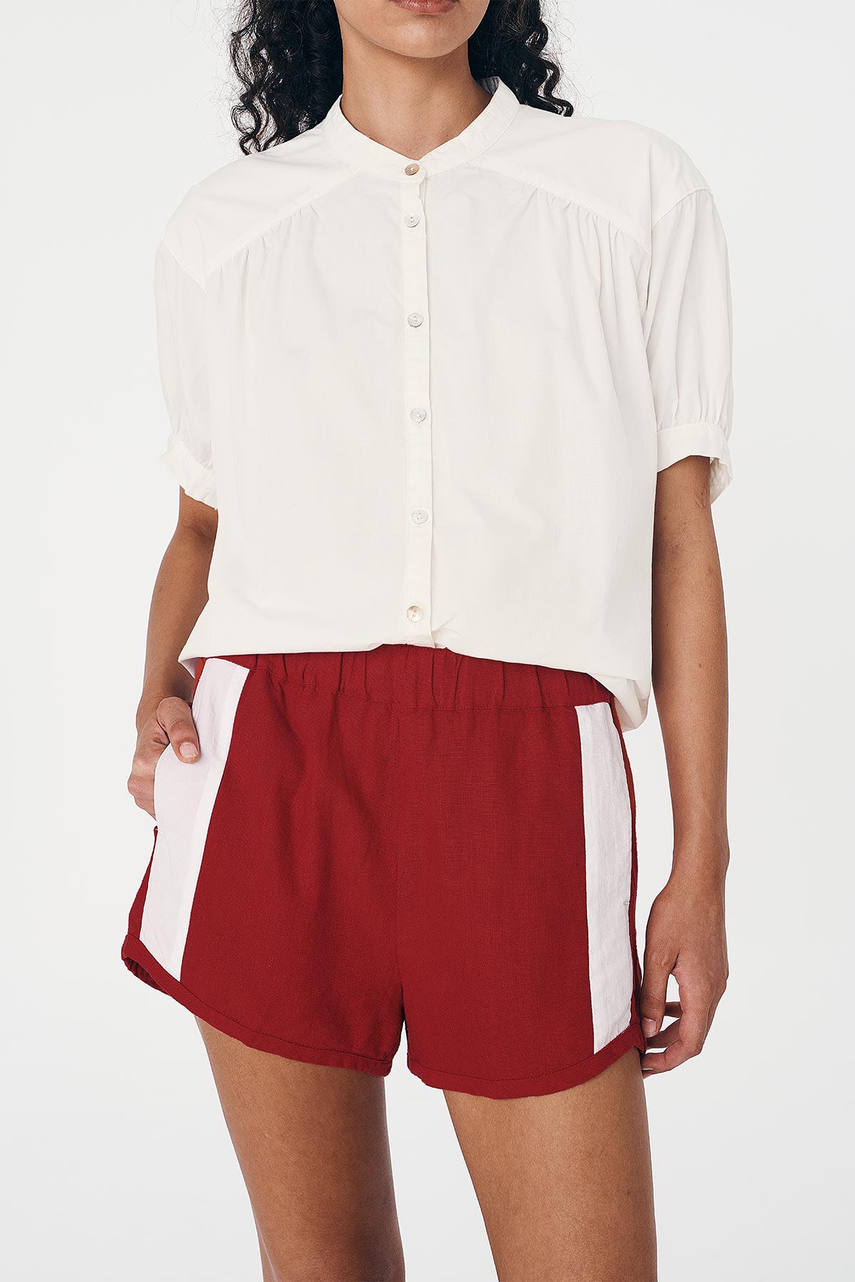 Person wearing a white button-up Cora Spring Blouse and red linen shorts with white block stripes. 